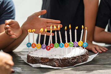 A birthday cake with white lit candles and colorful letters spelling “Happy Birthday.” The cake is dusted with powdered sugar and sits on a wooden table. Children eagerly surround the cake