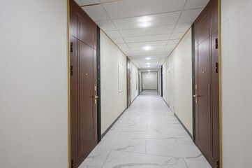 long corridor with brown doors on both sides. The floor is tiled with white marble-like tiles. The walls are white. Ceiling tiles with recessed lights