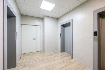modern corridor with light wood floors, two elevators, a white door, and tiled walls. Bright...