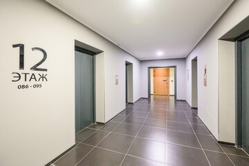 brightly lit hallway features dark tile flooring, gray walls, and a white ceiling. Elevator doors and a wooden door are visible