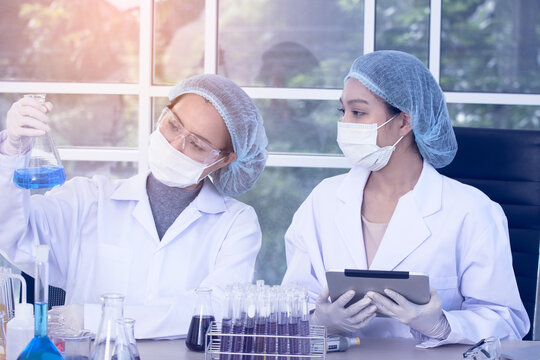 Two female scientists in lab coats and protective gear are performing an experiment, holding a flask with blue liquid while observing and taking notes on a tablet.