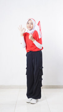 Indonesia independence day celebration with beautiful Asian girl wearing red and white outfit holding mini Indonesia flag decoration and showing OK hand gesture sign isolated on white background
