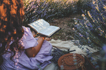 Girl in lavender dress reading a Bible in blooming lavender field. Peaceful summer moment with woven bag, straw hat, and wildflowers. Spiritual and tranquil atmosphere
