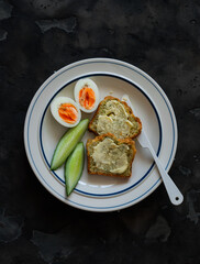Delicious breakfast - toasts with butter, boiled egg, fresh cucumber on a plate on a dark background, top view