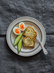 Delicious breakfast - toasts with butter, boiled egg, fresh cucumber on a plate on a grey background, top view