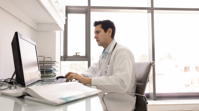 Focused young medical practitioner man working at computer in doctor office, typing on keyboard, writing electronic medical records, examination reports, using modern technology