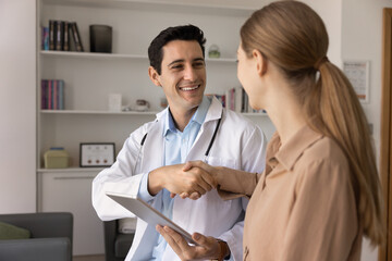 Positive confident handsome male doctor shaking hands with healthy young female patient, giving consultation with good healthcare news, perfect examination results, smiling