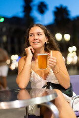 Woman with a glass of beer is sitting on the street near trendy bars, she is having a good time and waiting for friends. European woman hangs out in the evening in Barcelona