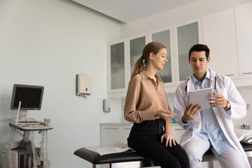 Positive young Latin practitioner pan showing ultrasound electronic image on tablet to female patient, sitting on medical couch, explaining diagnosis, examination result. Wide shot with copy space
