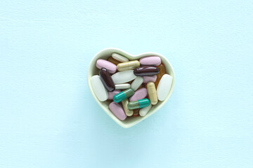 Green heart shaped bowl with various colored dietary supplement pills on light blue wooden background. Health supplement concept. Top view with copy space.