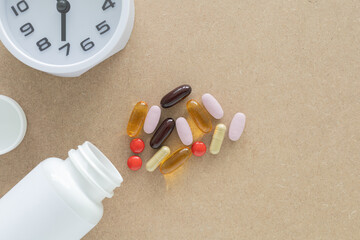 Top view, a group of scattered supplement pills and capsules and white plastic pill bottle, clock, on a brown background. Health, wellness, and care pharmacology concept.