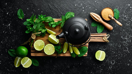 Mint tea with lime: fresh green limes, mint, brown sugar and a kettle of boiling water. On a black stone background. Top view.