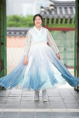 Fototapeta premium In June in Seoul, a Korean woman in her twenties stands at the traditional gate of Gyeongbokgung Palace, wearing a soft sky-blue hanbok and gazing amid ornate palace grounds under a clear summer sky.