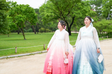 Fototapeta premium Two Korean women in their twenties stroll through a lush green park at Gyeongbokgung Palace, wearing cute pink and sky-blue hanbok, smiling and chatting beneath a clear summer sky. Seoul, June.