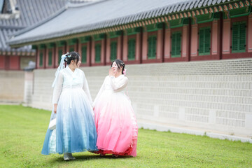 Two Korean women in their twenties walk across a grassy lawn before a historic hall at Gyeongbokgung Palace, wearing cute pink and sky-blue hanbok under a clear summer sky peacefully. Seoul, June.
