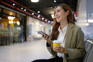 Smiling businesswoman using smartphone and holding coffee cup while sitting in a modern indoor space.