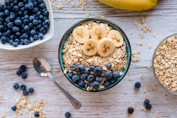 Top view of a porridge oatmeal, healthy breakfast bowl with oats, banana slices, and blueberries on a rustic wooden table, surrounded by fresh fruit and scattered oats.