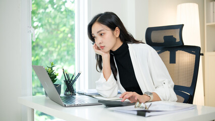 Asian businesswoman working on laptop and using calculator, appearing thoughtful at modern office desk.