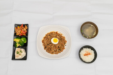 A Thai basil minced pork set meal arranged on a tray, including a portion of stir-fried spicy basil pork, a bowl of steamed white rice, three side vegetables, and a bowl of soup. The presentation refl