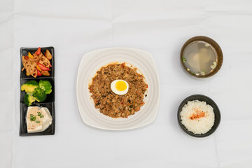 A Thai basil minced pork set meal arranged on a tray, including a portion of stir-fried spicy basil pork, a bowl of steamed white rice, three side vegetables, and a bowl of soup. The presentation refl