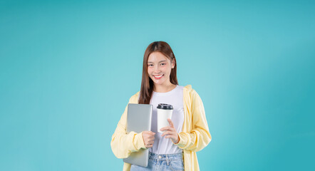 Fototapeta premium Young Asian woman smiling while holding a laptop and coffee cup against a sky blue background, representing modern lifestyle, online learning, remote work, and technology.