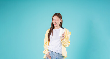 Happy young Asian woman holding a coffee cup while smiling against a blue background, symbolizing...