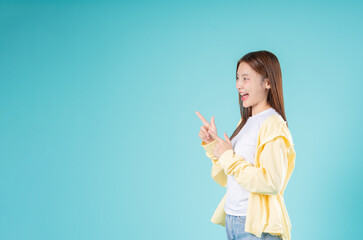 Excited young Asian woman pointing to the side with both hands against a blue background, ideal for...