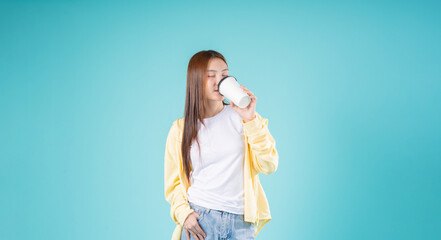 Relaxed young Asian woman enjoying the aroma of coffee while standing against a blue background,...