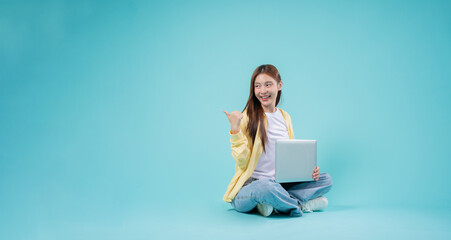 Smiling young Asian woman sitting cross-legged with a laptop, pointing to the side against a blue...