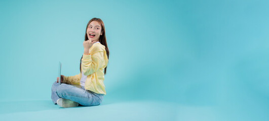 Cheerful young Asian woman sitting cross-legged with a laptop, pointing to the side against a...