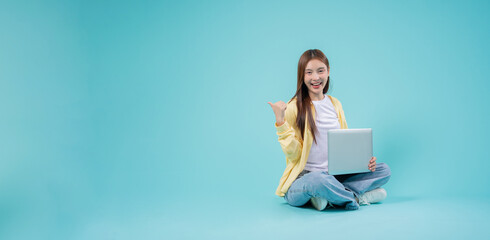 Smiling young Asian woman sitting cross-legged with a laptop, pointing to the side against a blue...