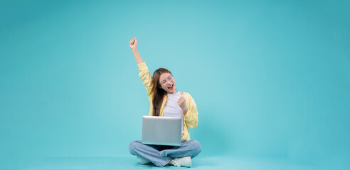 Excited young Asian woman sitting cross-legged with a laptop, raising her hand in celebration...