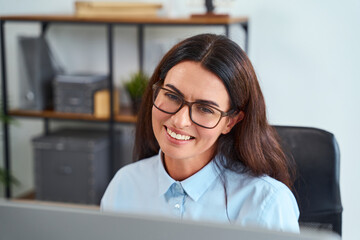 Confident Woman Wearing Glasses Smiling at Computer While Working in a Modern Office Environment,...