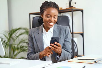 Professional Woman In Office Suit Smiling While Using Smartphone. Bright Workspace Background...