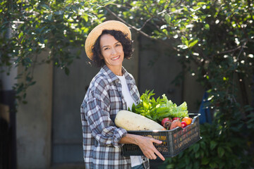 Happy  middle aged woman wearing a straw hat holds a box of vegetables picked in her garden.. 