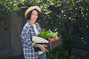 Happy  middle aged woman wearing a straw hat holds a box of vegetables picked in her garden.. 