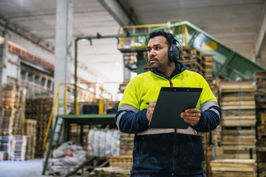 Supervisor wearing ear protection and high visibility jacket writing notes on clipboard in a wooden pallet recycling factory