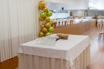 Support table in a decorated banquet hall. Empty glasses and a bread basket neatly arranged for guests, with festive gold and green balloons creating an inviting detail.