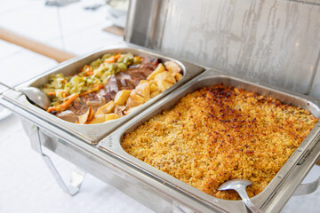 Buffet lunch served at a catered event. Hot chafing dishes feature a meat dish with roasted potatoes and vegetables, and a baked fish dish. Ideal for gatherings.