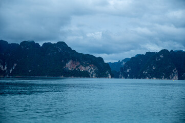 Stunning limestone cliffs covered with lush greenery stand tall against a tranquil lake under cloudy skies in southern Thailand.