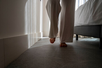 A person is walking barefoot in a room with a bed in the background. The room is dimly lit, and the person's shadow is cast on the floor