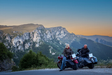 Couple sitting on motorcycles enjoying a quiet moment at sunset in the mountains, surrounded by dramatic cliffs and natural beauty.