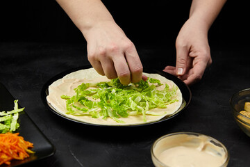 Hands of a person preparing a fresh wrap by adding crisp lettuce on a tortilla, surrounded by colorful vegetables and sauces, showcasing healthy meal preparation