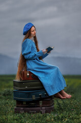 Dreamy vintage girl in blue dress reading a book on stacked suitcases in countryside field under cloudy sky
