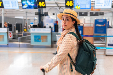 Mid shot of woman in an elegant suit, straw hat and backpack stands near the check-in windows in...