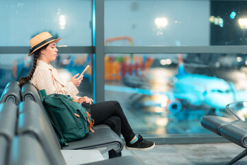 Wide shot of young Caucasian woman in a hat and jacket using smartphone in airport waiting area sitting in chair. Web banner of travel with copy space. Concept of boarding at terminal before flight