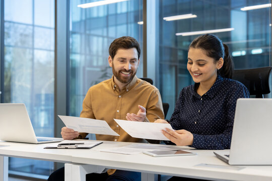 Two colleagues review documents at a modern office desk, appearing engaged and collaborative.
