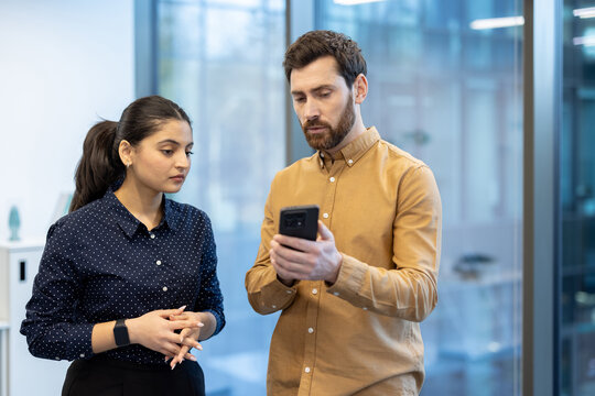 A man shows something on his phone to a woman in an office setting, with a focus on their interaction. - Powered by Adobe