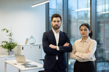 A professional business team, a man and a woman, stand confidently in an office setting, arms crossed, looking directly at the camera.