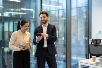Two professionals in business attire are looking at a smartphone, smiling, within a modern office setting, near large windows.